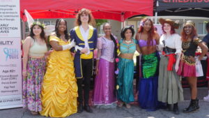 Photo of Brown Beauty Magic group. They are dressed as princesses Tinkerbelle, Belle, the Beast, Tangled, Jasmine, Ariel, and a pirate.