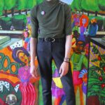 Photo of a Hispanic man with curly dark hair standing in front of a backdrop. He is wearing a gray short sleeve turtleneck, black trouser pants and gray suede shoes.
