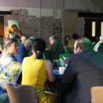 Photo of a table of people talking about something very interesting, as they are all leaning in to the animated speaker. a woman in yellow with black hair with her back to the camera.