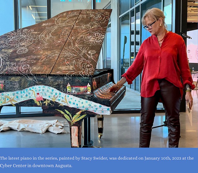 Staci Swider with her painted piano at the Jessye Norman School of Arts. Staci is a Caucasian woman with blonde/gray hair wearing a red button down blouse and black pants.