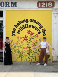 Galadra and Heather posing in front of an Augusta mural. They are both Caucasian individuals with long brown hair, and the mural is bright yellow with colorful wildflowers