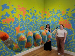 Galadra and Heather posing in front of the micro gallery at the Westobou. The walls are a bright green covered in blue and bright orange speckles.