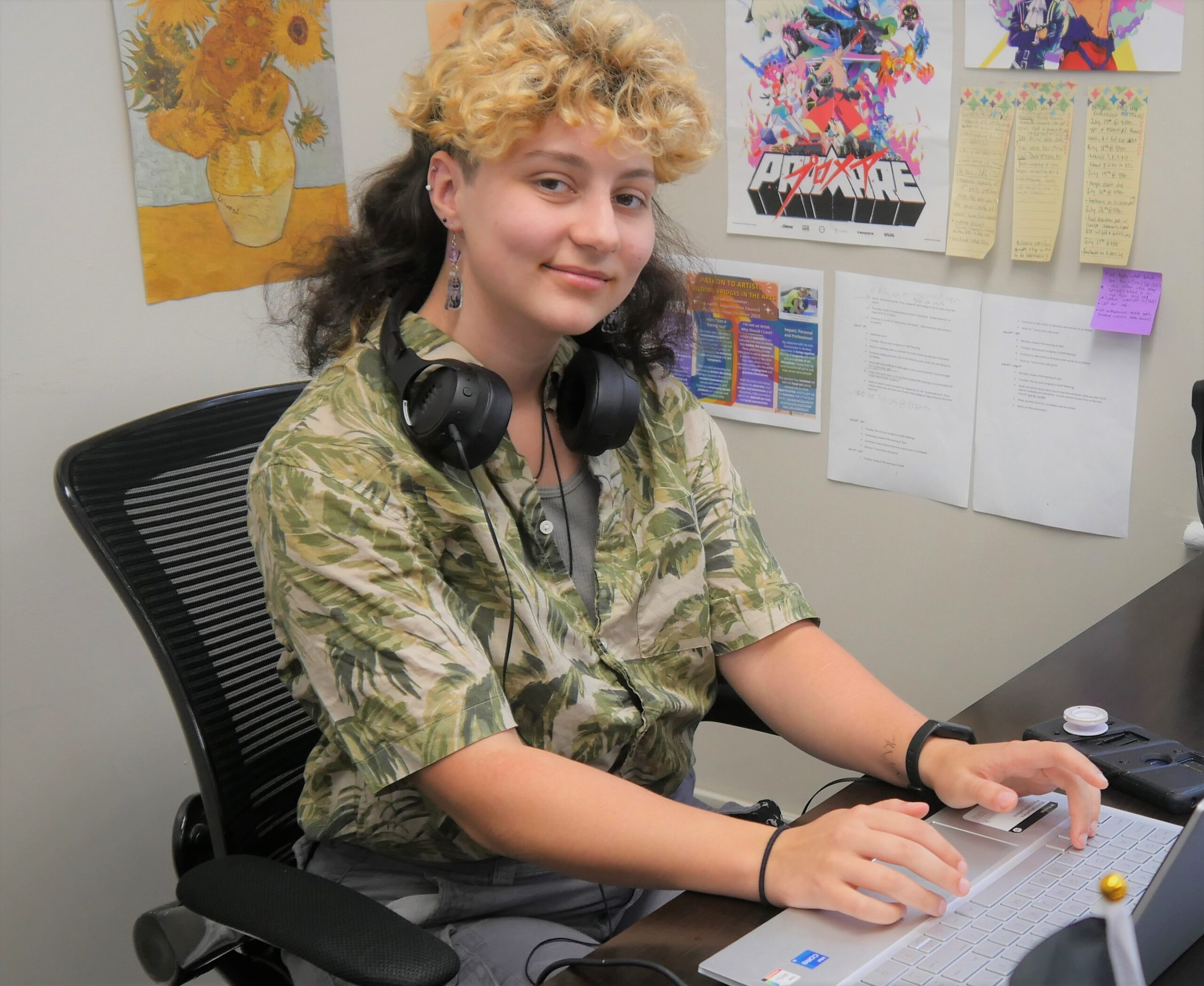 Galadra Plummer sitting at a desk. They are a caucasian person with a brown and blonde mullet and a green patterned shirt