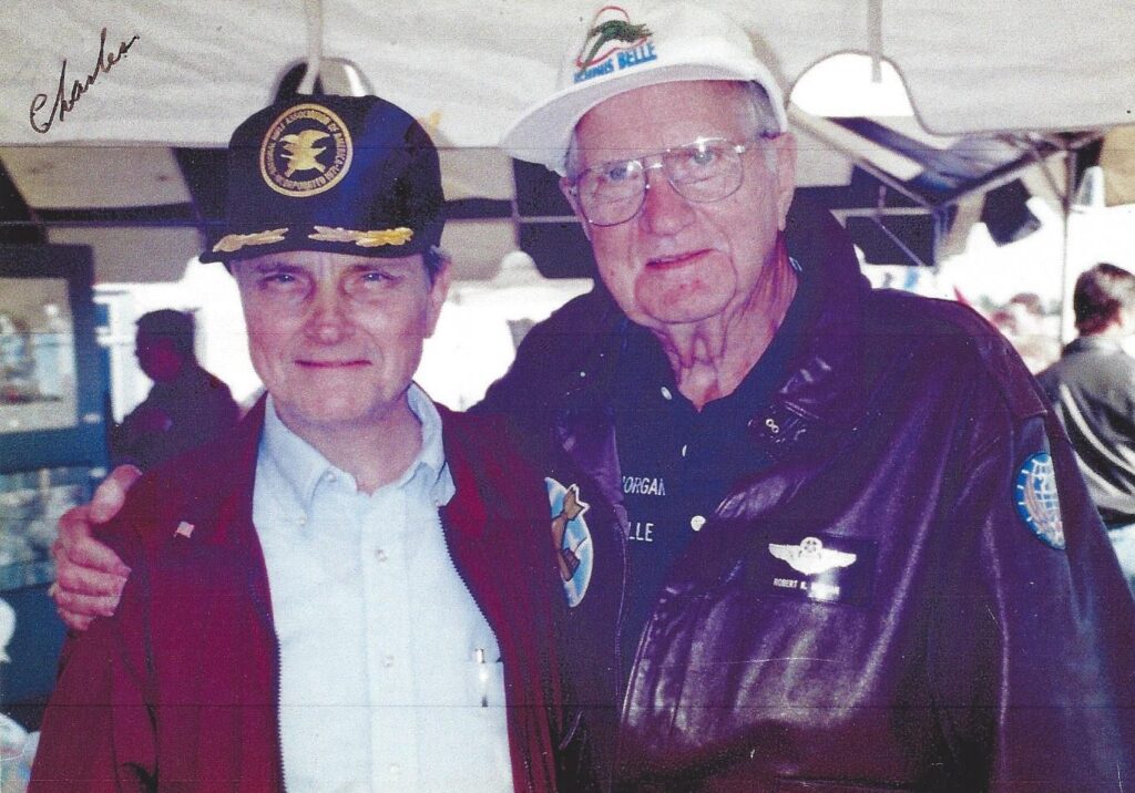 Two older men wearing captains wings and has. The man on the left is wearing a red jacket. The man on the right is in a black leather jacket. Both are Caucasian.