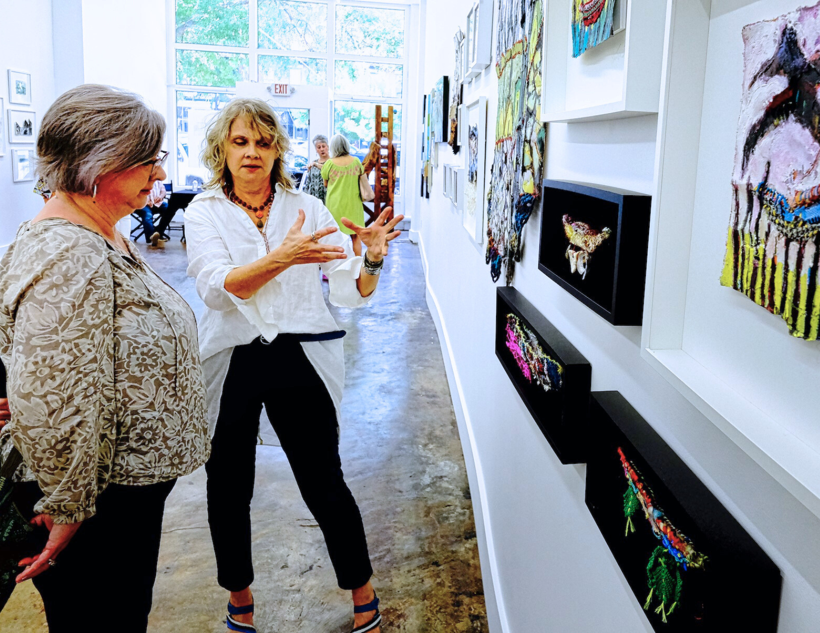 photo of Staci Swider talking to art patron. Staci is a Caucasian woman with short blonde hair. She is wearing a white button down shirt with black skinny jeans.