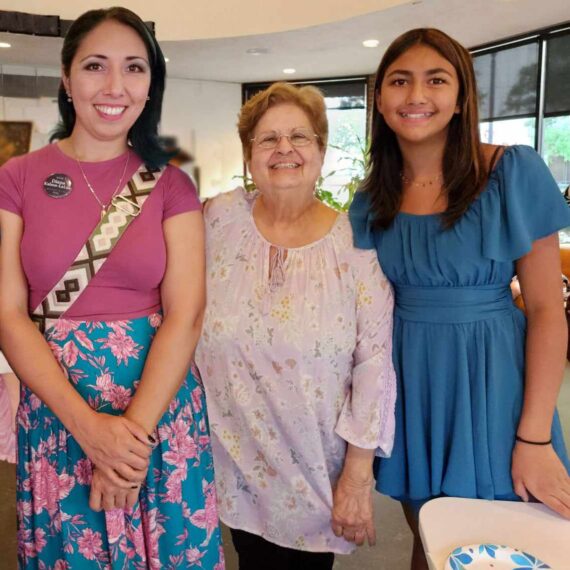 Three generations of yarn artists. Grandmother, Daughter and Granddaughter all stand together. The grandmother, who is standing at the center, has short graying hair and is wearing a white shirt, the Daughter on the Left has dark brown hair and is wearing a pink shirt, the Granddaughter has brown hair and is wearing a blue dress.