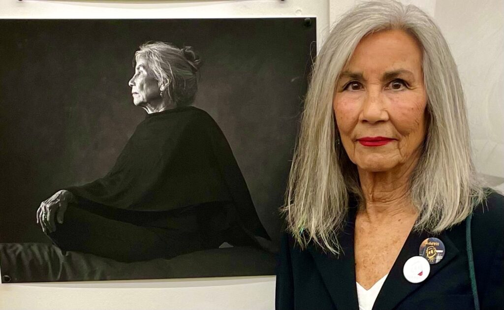 Gerry O'Meara standing in front of a portrait of herself. She has long silver hair and is wearing a black cardigan with a white rose broach.