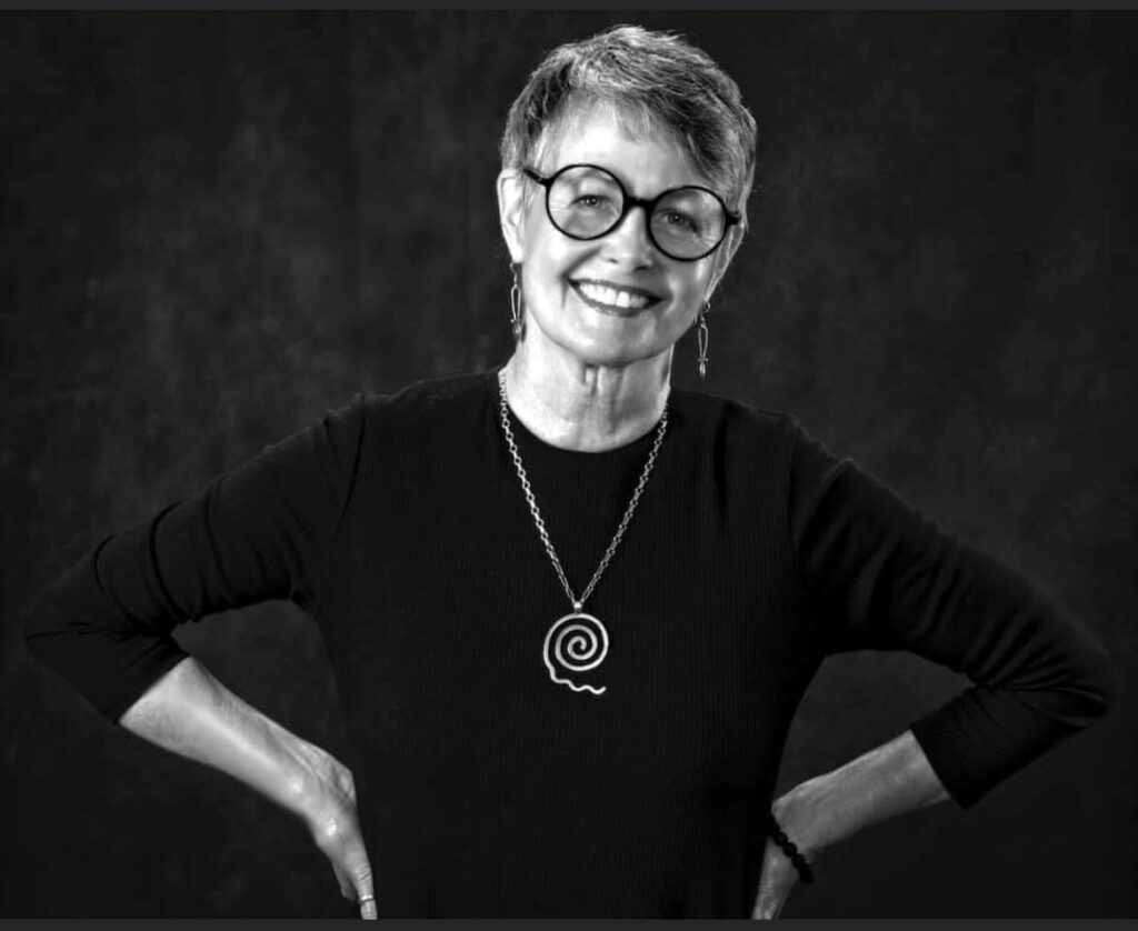 Black and White photo of a woman wearing a black shirt, necklace and glasses. She has short hair and is smiling with her hands on her hips.