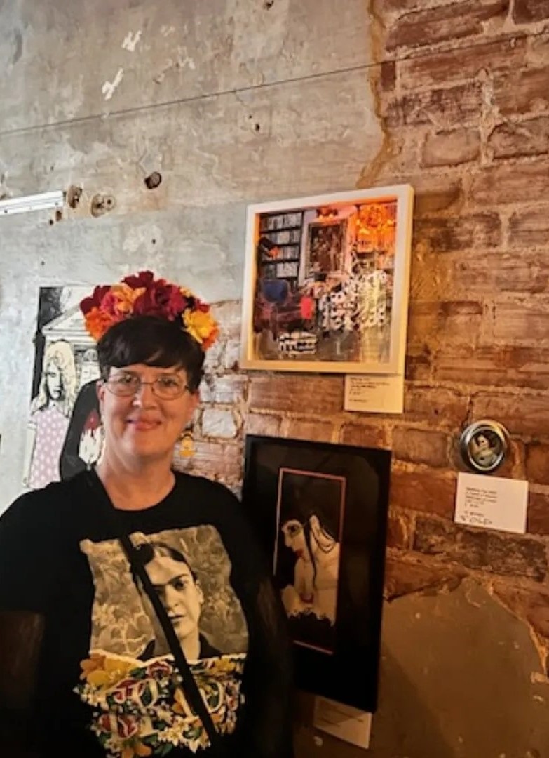 Photo of woman standing infront of artwork. She has short brown hair and is wearing a flower crown.