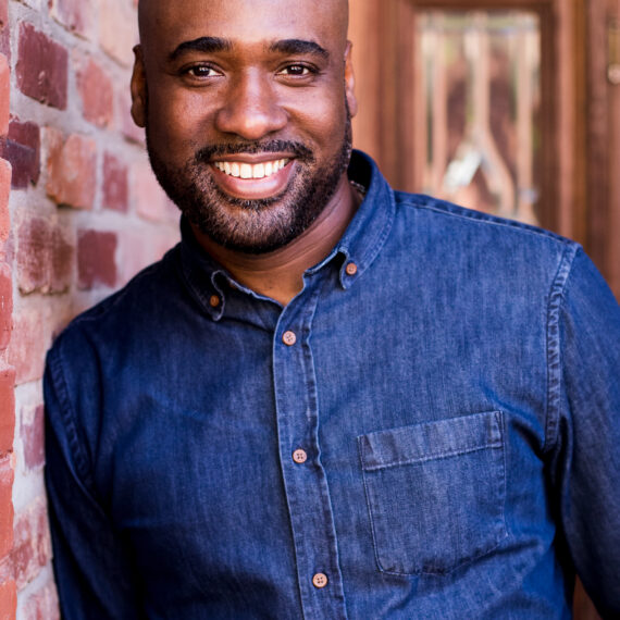 Portrait of Ray Sturkey. He is an African American man with facial hair. In the photo he is smiling, wearing a denim button down. He is standing by a red brick wall.
