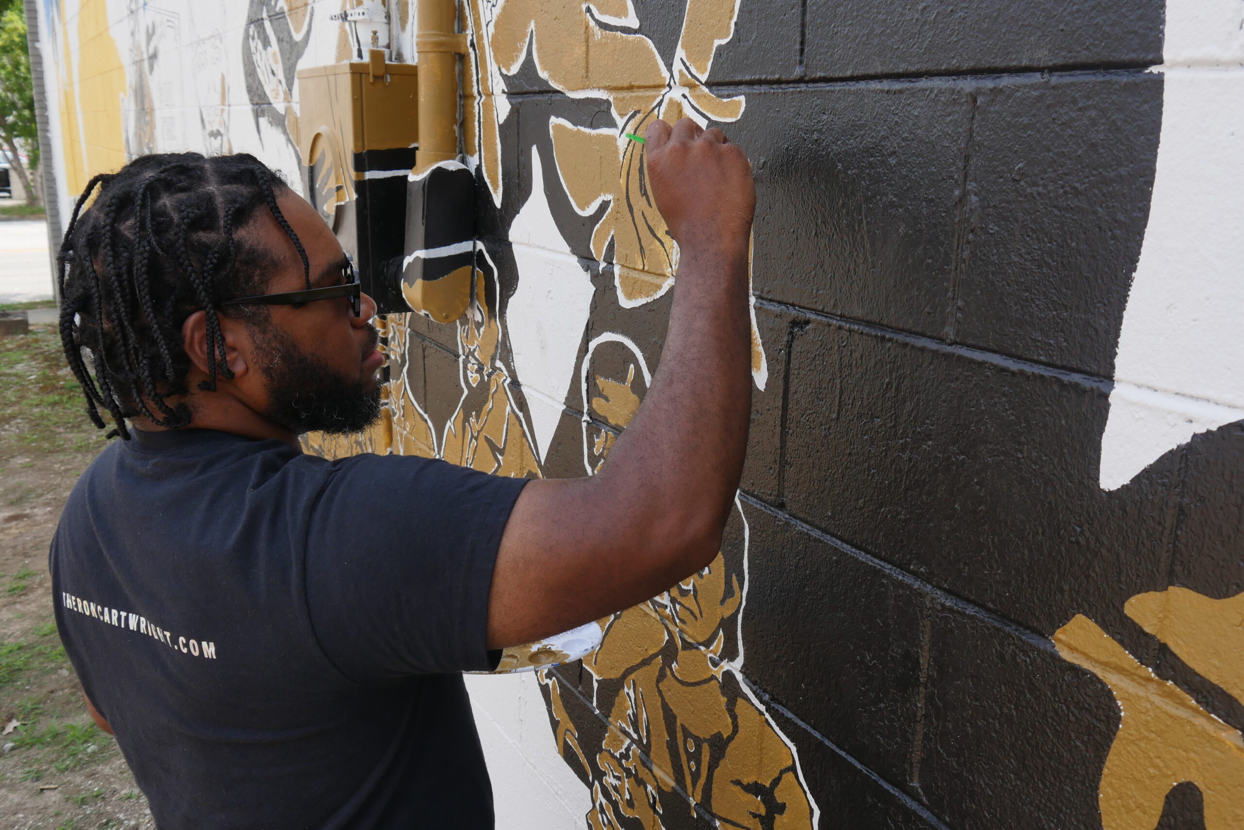 African American man painting a mural. He has short dreaded hair and is wearing sunglasses, a black graphic t-shirt and jeans.
