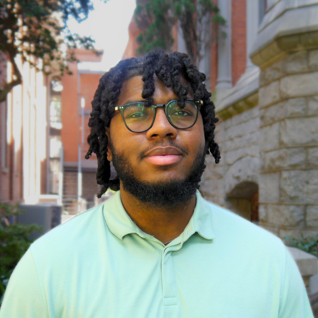 Man standing in front of a brick building. He is African American with short hair, black rimmed glasses and a beard. He is wearing a light green polo shirt.