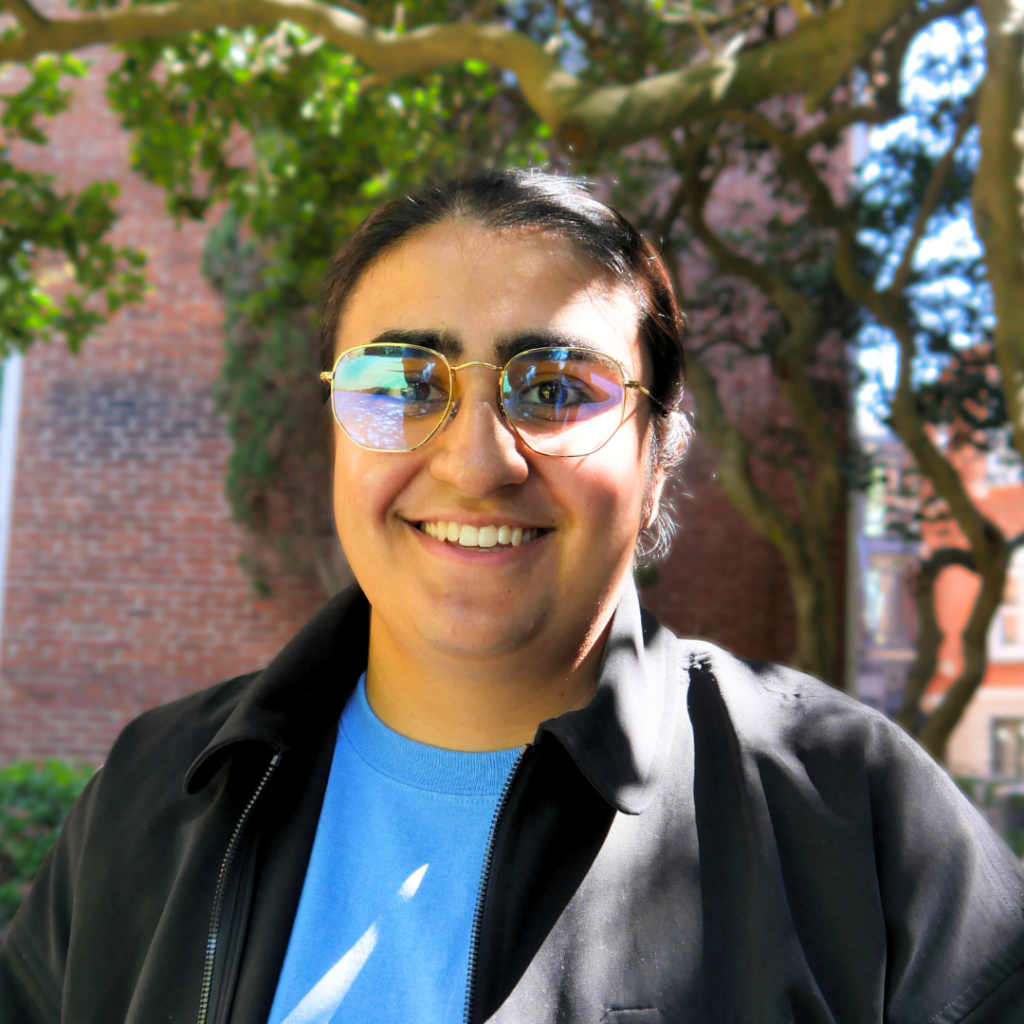 A woman standing in front of a brick building. She is of Indian descent. She has her dark hair pulled back into a pony tail, is wearing gold rimmed glasses and a blue t-shirt with a black jacket.