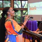 A African American woman singing into a microphone as someone plays piano behind her.
