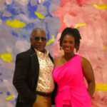 A African American father and daughter posing in front of a painted backdrop. The man is wearing a black coat and the woman is wearing a hot pink dress.