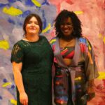 A Caucasian woman and a African American woman posing in front of a backdrop
