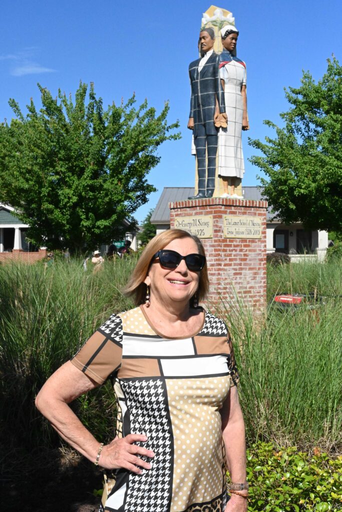 Caucasian woman standing in front of a sculpture. She is smiling, wearing sunglasses and a patchwork dress.