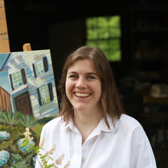 Photo of a Caucasian woman. she has shoulder length brown hair. She is smiling with a painting of a house. She is wearing a white button down shirt.