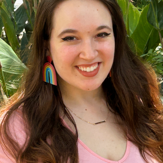 Caucasian woman with long brown hair. She is wearing a pink shirt and rainbow earrings.