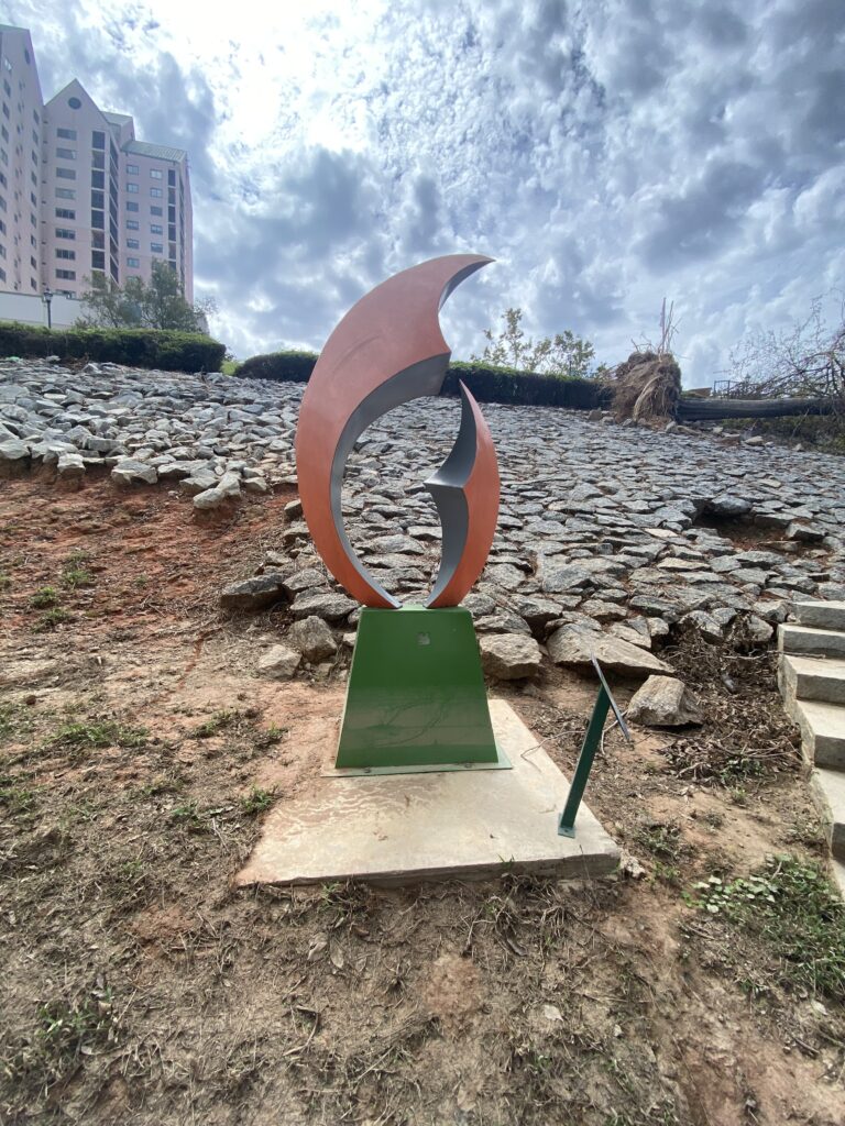 Photo og a metal sculpture that is painted orange and green. It is in the shape of a flame. In the background there is a stone laid hill that leads to a path, hotel and office building.