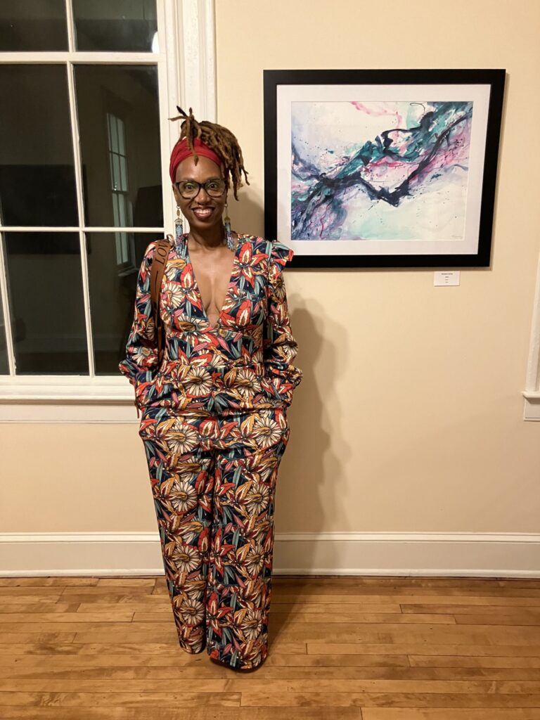 African American woman at an art show. She is dressed in a colorful romper outfit with a red head scarf. She has short dreaded hair that is put up. She is standing in front of a colorful, abstract watercolor painting.