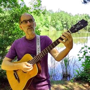 A caucasian man in a purple t-shirt playing acoustic guitar near a lake.