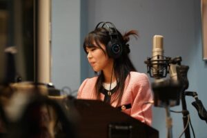An Asian woman seated at a piano. She is wearing headphones and playing music. there is a microphone near her.