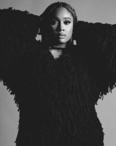 An African American woman dressed in all black with her hands behind her head.