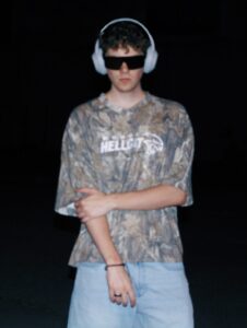 a caucasian man wearing a camoflauged t-shirt and jeans with dark glasses and a pair of white headphones.