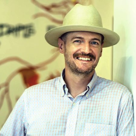 Matt Porter is a Caucasian man with a brown beard and mustache. He is wearing a white, wide brimmed hat with a blue button down shirt.